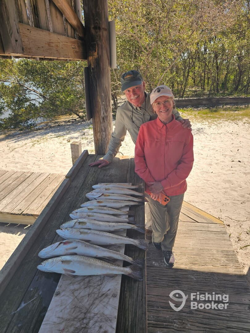 Two anglers proudly posing with a catch of speckled trout displayed on a board at a scenic fishing spot.