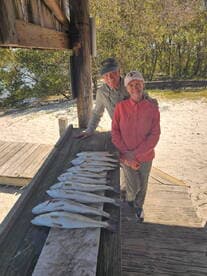 Two anglers proudly posing with a catch of speckled trout displayed on a board at a scenic fishing spot.