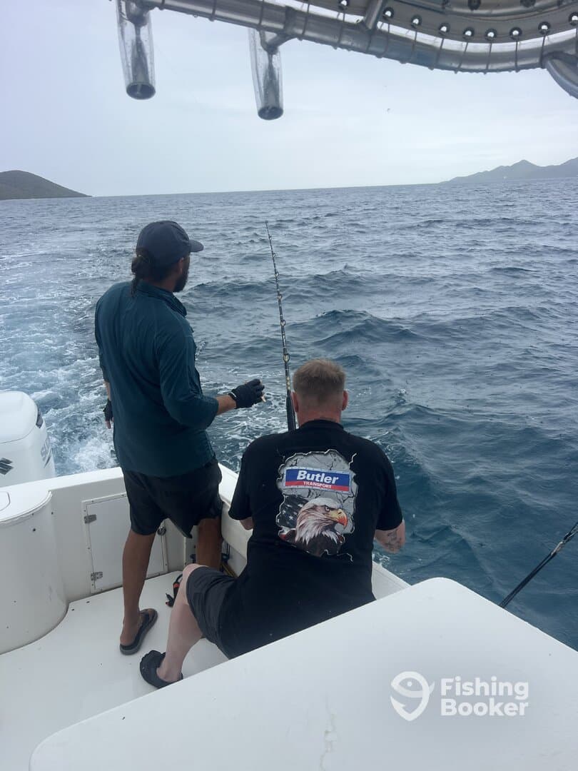 Two anglers actively fishing on a boat, with one reeling in a line while the other assists, surrounded by open water and distant hills.