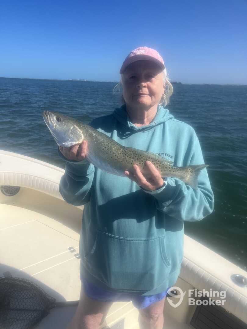 An angler proudly displaying a large Speckled Trout while fishing on a boat in calm waters.