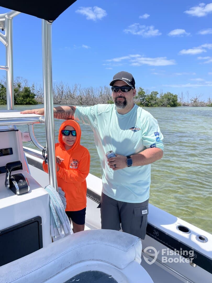 A father and son enjoying a day on the water, with the son wearing an orange hoodie and sunglasses, while the father holds a drink on a fishing boat.