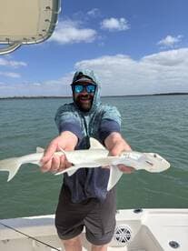 Angler proudly displaying a small shark while fishing on a boat in calm waters.