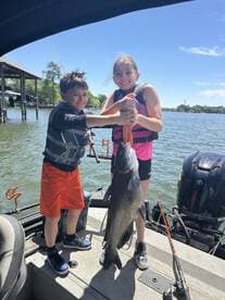 Two young anglers proudly displaying a large catfish while fishing from a boat on a sunny day.