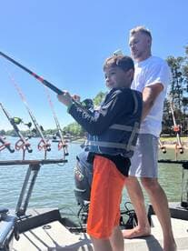 A young angler, wearing a life jacket, is joyfully reeling in a catch while fishing with an adult on a sunny day.