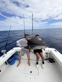 Two young anglers proudly displaying a large Sailfish aboard a fishing boat in open waters.