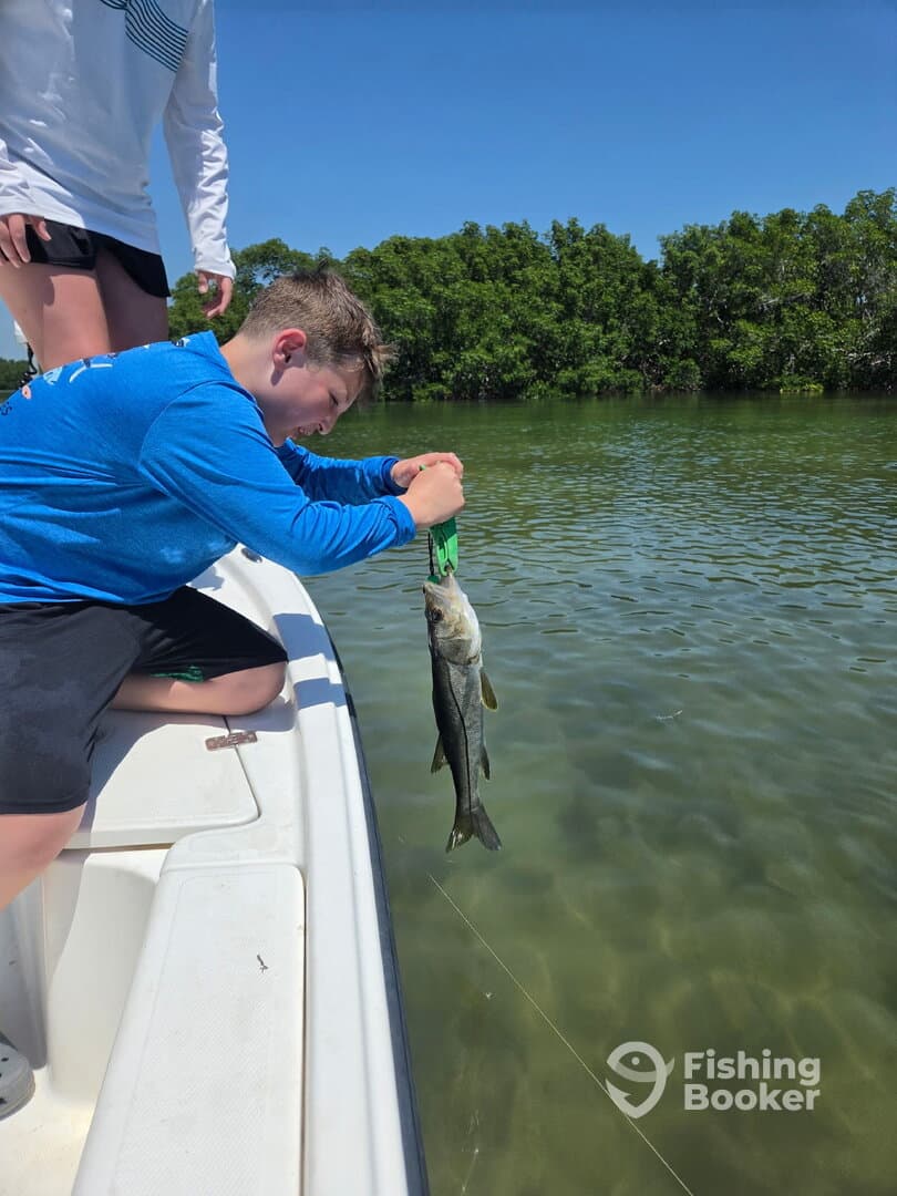 A young angler is reeling in a Snook while fishing from a boat in a scenic coastal environment.