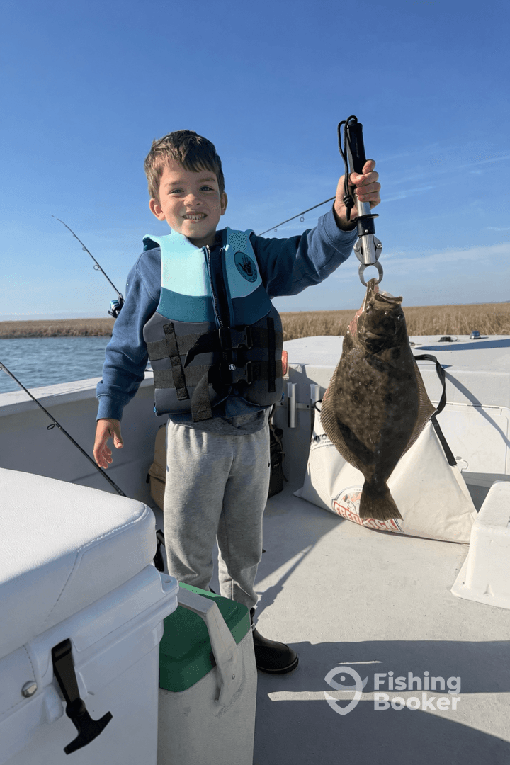 A young angler proudly holding a Flounder while fishing on a boat, showcasing a successful catch in a scenic coastal environment.