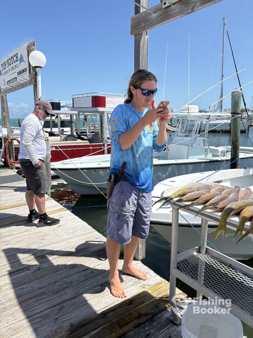 A young man is standing on the dock, focused on his phone, with boats in the background. No fish are prominently displayed.