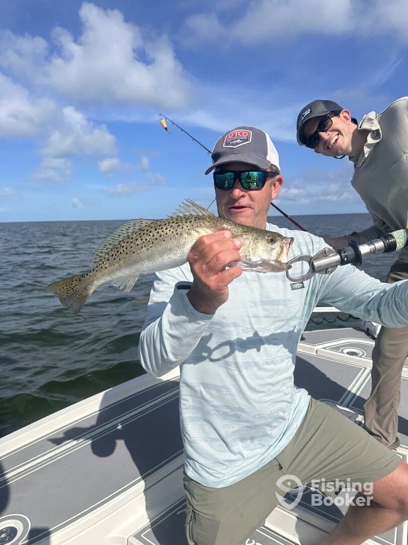 Angler proudly displaying a Speckled Trout while fishing on a boat, with a sunny sky and calm waters in the background.