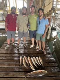 A group of four friends proudly displaying their catch of Redfish on a dock after a successful fishing trip.