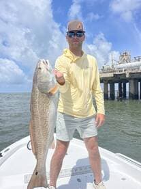 Angler proudly displaying a large Redfish while standing on a boat, with a scenic backdrop of clouds and a pier.