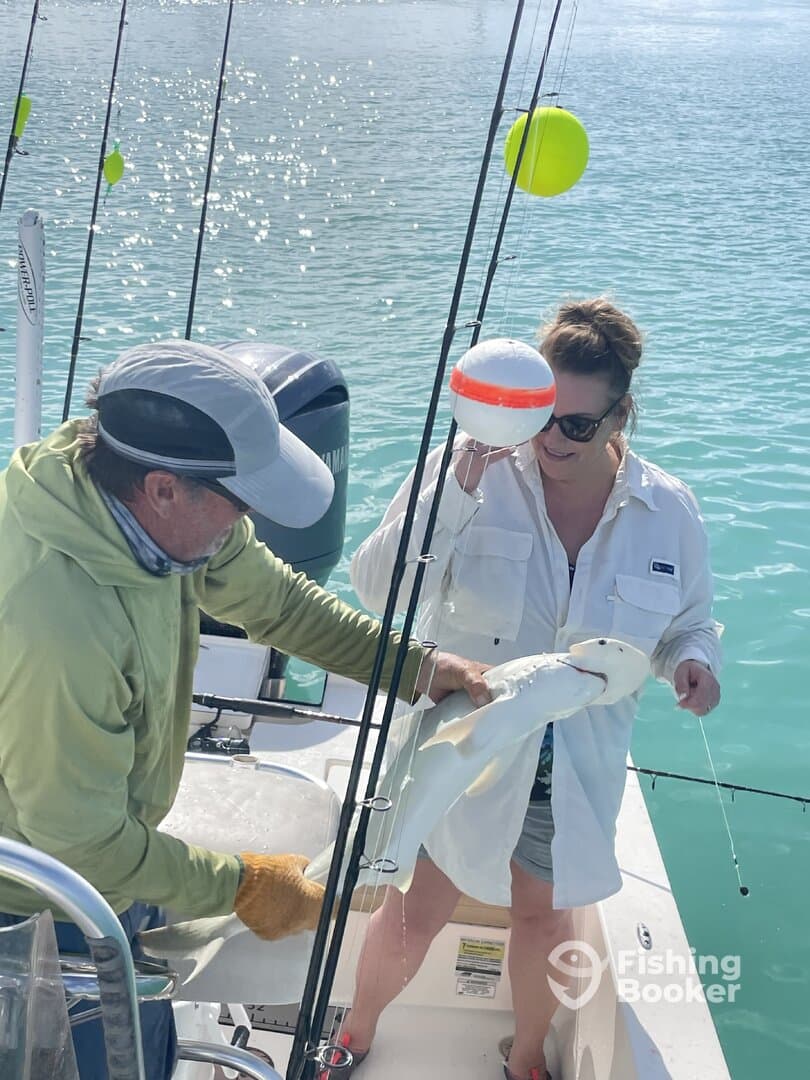Two anglers on a boat are actively engaged in fishing, with one holding a freshly caught fish while the other assists. The scene captures the excitement of fishing in clear blue waters.