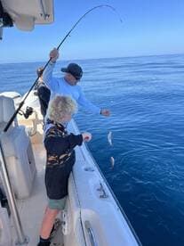 A young angler and an adult are actively fishing on a boat, with multiple fish being reeled in during a sunny day on the water.