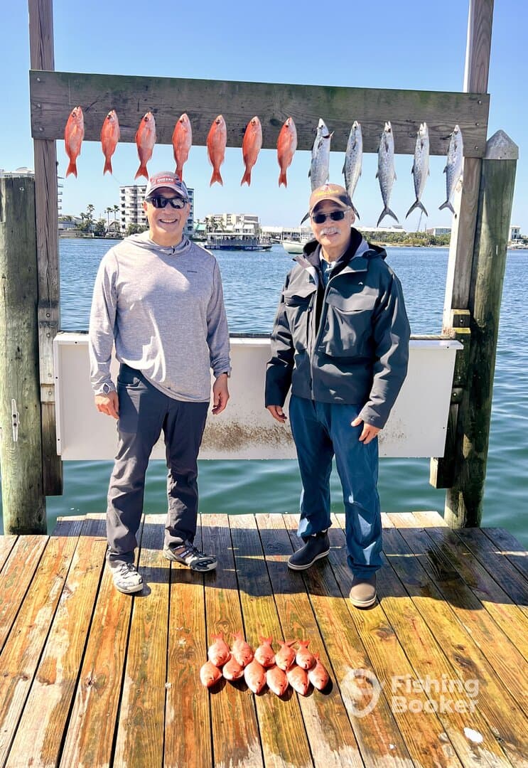 Two anglers proudly standing in front of a catch board displaying several Red Snapper and Mackerel after a successful fishing trip.