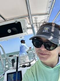 A woman takes a selfie while a man is casting a line in the background, showcasing an exciting fishing trip on a boat.