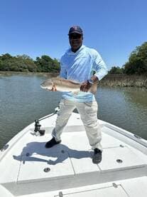 Angler proudly displaying a large Redfish while standing on the bow of a boat in a scenic fishing environment.