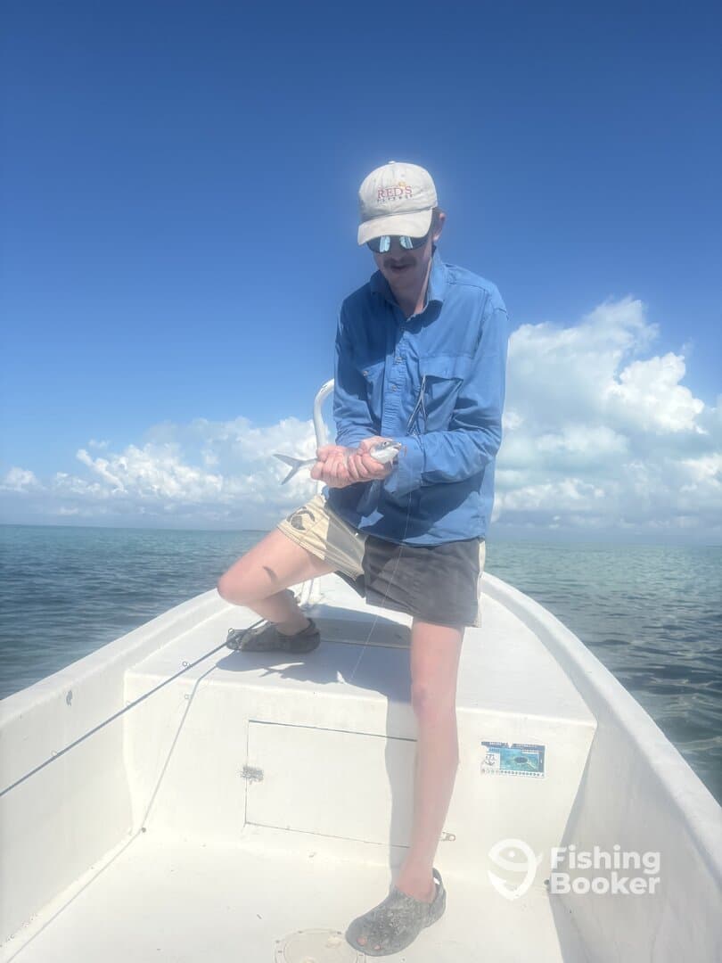 A young angler is actively reeling in a fish while standing on the bow of a boat, surrounded by clear blue waters and a bright sky.
