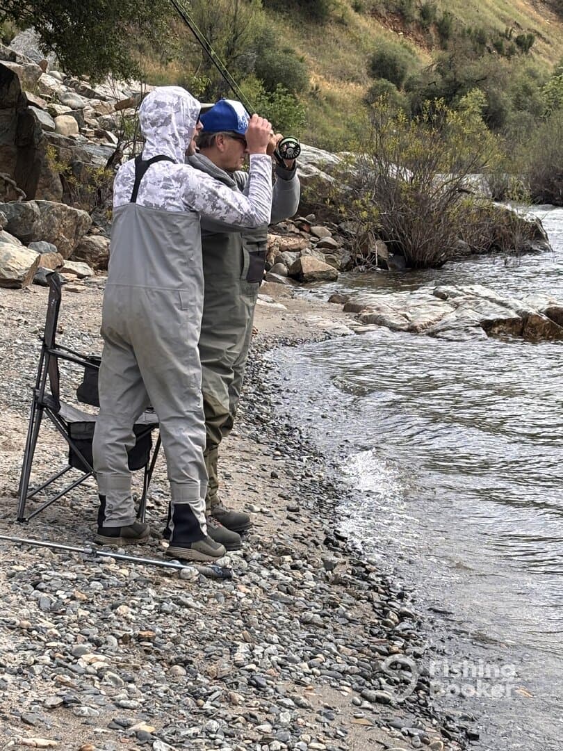 Two anglers are actively fishing along a rocky shoreline, one casting while the other observes the water, showcasing a serene fishing environment.