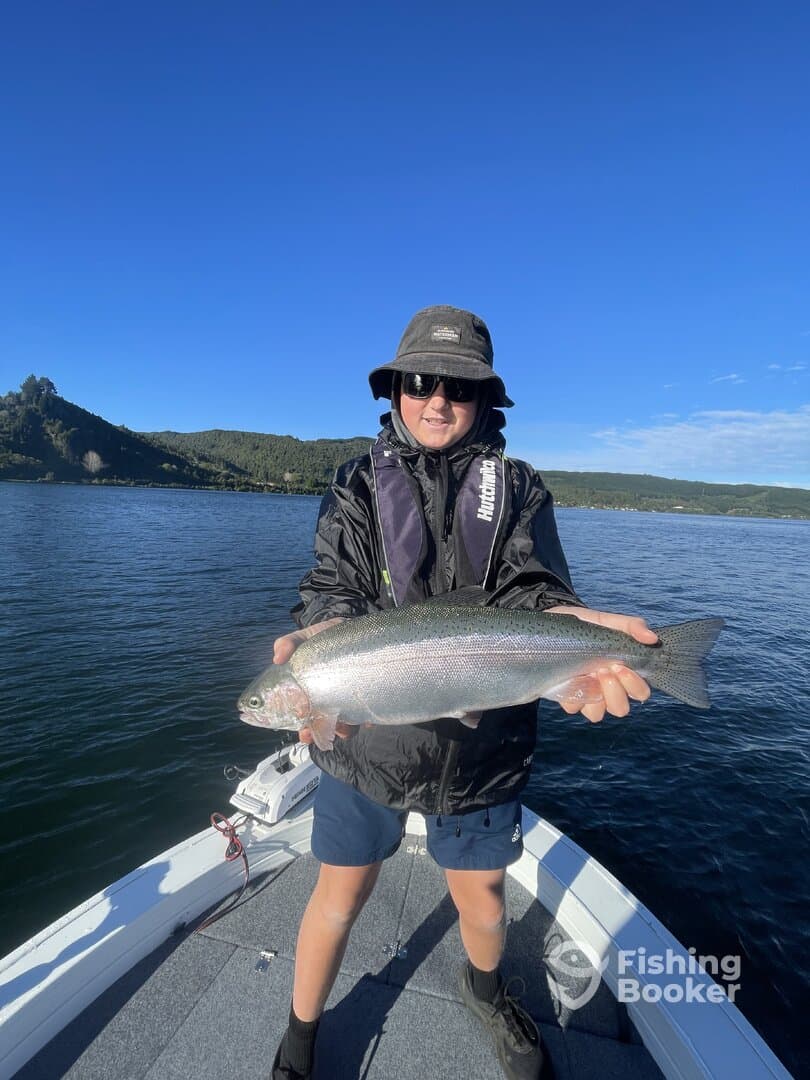 Young angler proudly displaying a Rainbow Trout while fishing on a serene lake, showcasing a beautiful day on the water.