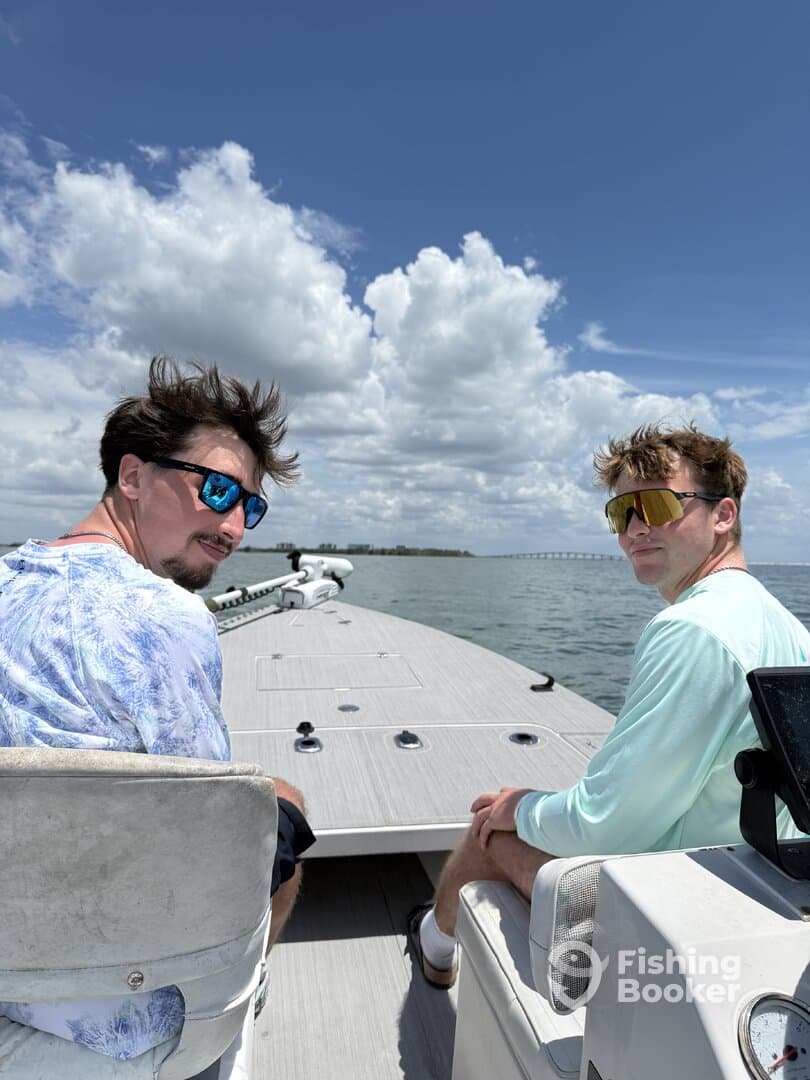 Two anglers enjoying a sunny day on a fishing boat, with a clear view of the water and clouds in the background.