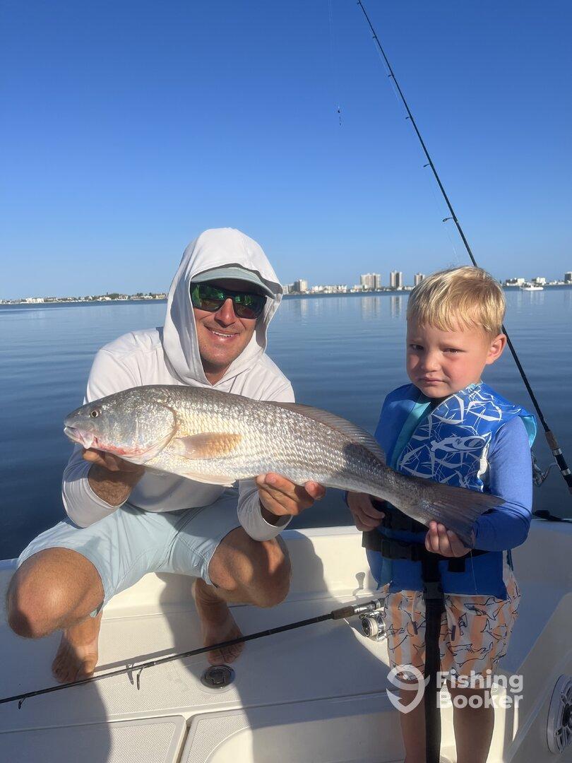 A proud young angler and an adult display a large Redfish while fishing on a calm day, showcasing a successful outing.