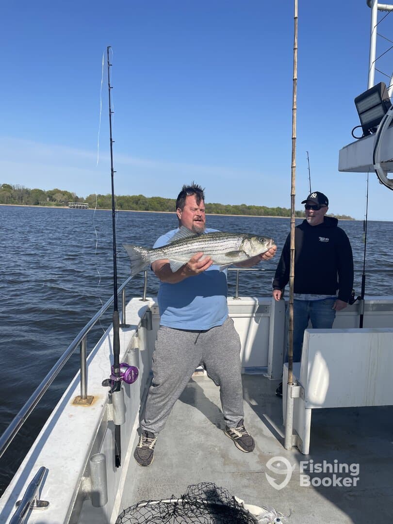 Angler proudly displaying a large Striped Bass while fishing on a charter boat, with another fisherman in the background.