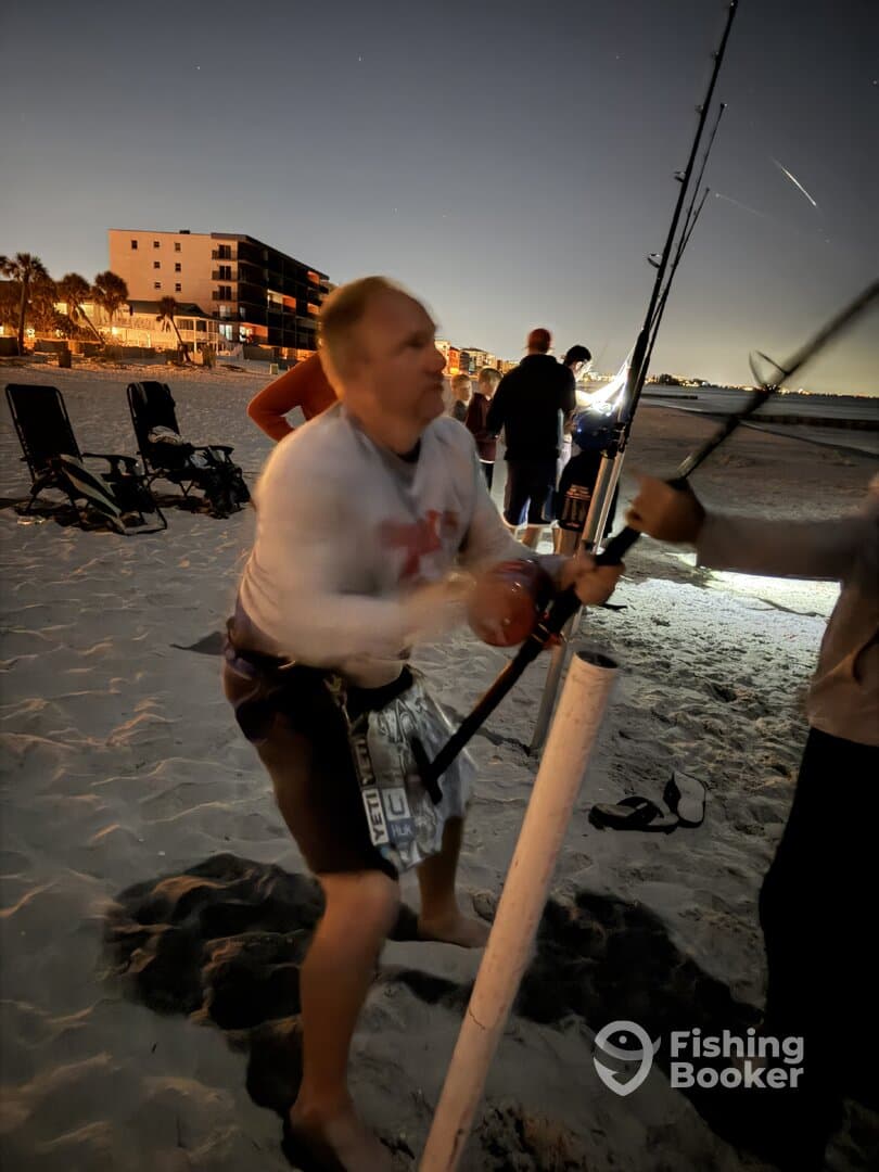 An angler actively reeling in a catch on the beach at night, surrounded by fellow fishers and illuminated by ambient lights.