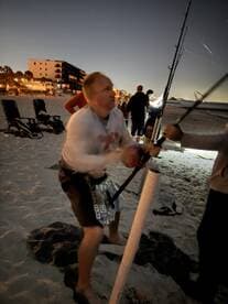 An angler actively reeling in a catch on the beach at night, surrounded by fellow fishers and illuminated by ambient lights.