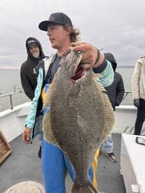 A young angler proudly displaying a large Halibut while fishing on a boat, with fellow anglers in the background.