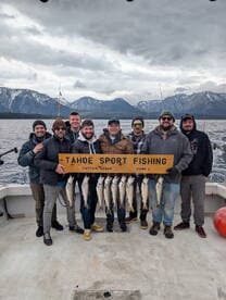 A group of anglers proudly displaying their catch of Lake Tahoe fish on a charter boat, with stunning mountain scenery in the background.