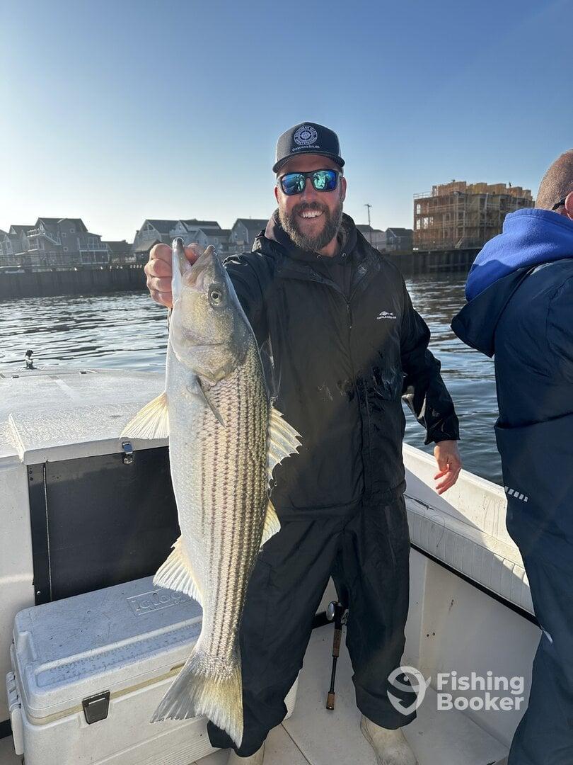 Angler proudly displaying a large striped bass while fishing from a boat, with a scenic waterfront in the background.