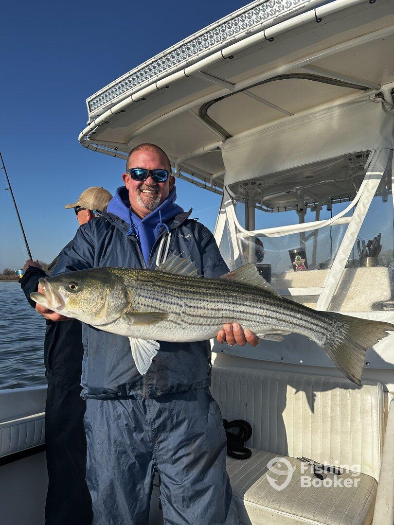 Angler proudly displaying a large Striped Bass while fishing on a boat, showcasing a successful day on the water.