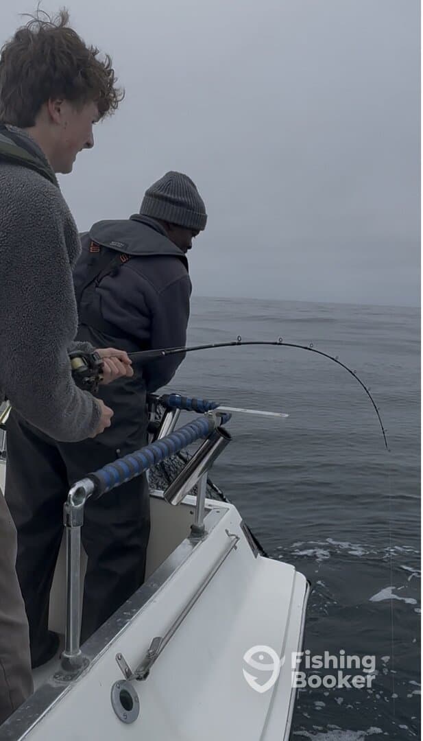 Two anglers engaged in reeling in a fish while fishing on a boat in overcast conditions.