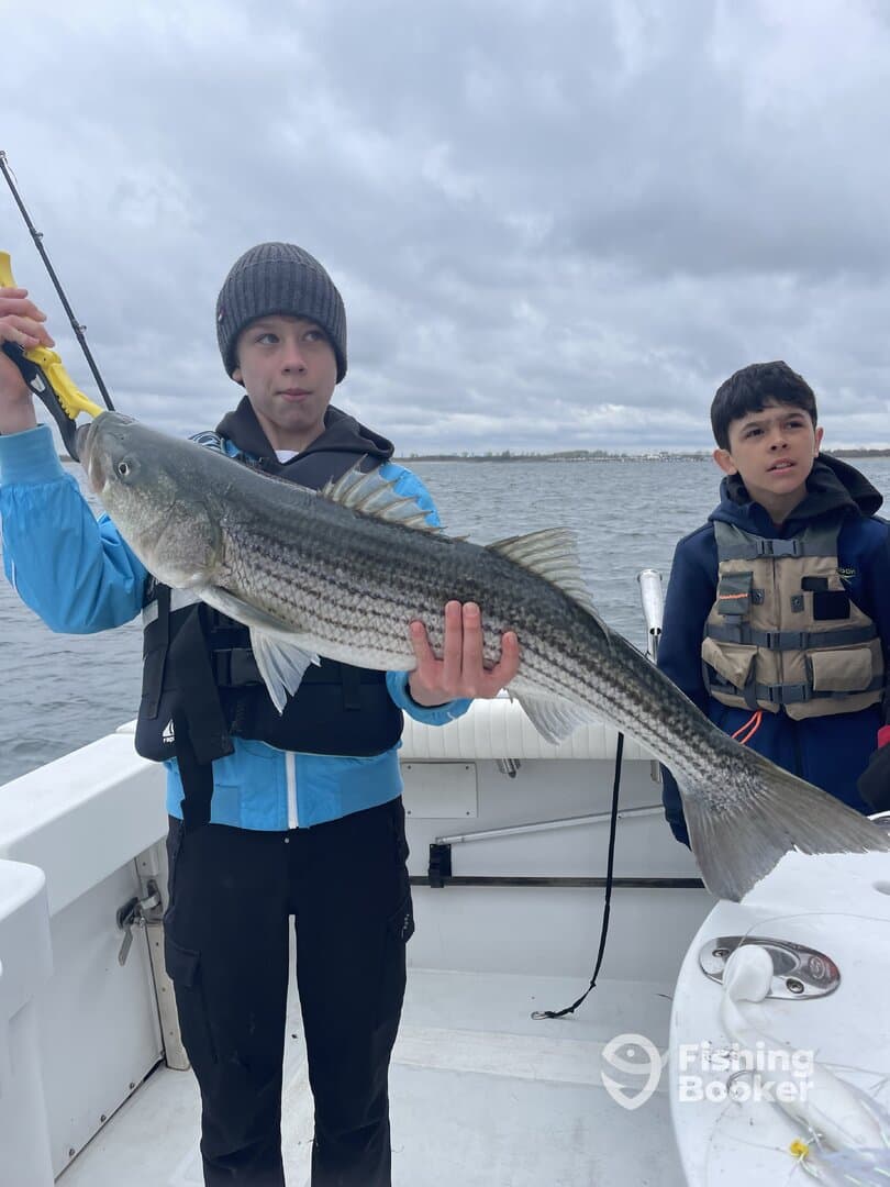 Two young anglers on a boat, with one proudly displaying a large Striped Bass caught during a fishing trip.