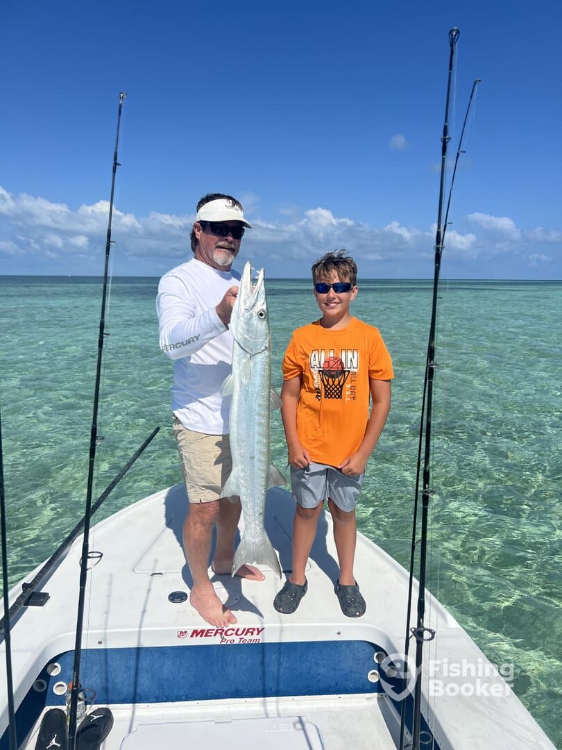 A young angler and an adult proudly displaying a large Barracuda on a boat in clear waters, showcasing a successful fishing trip.