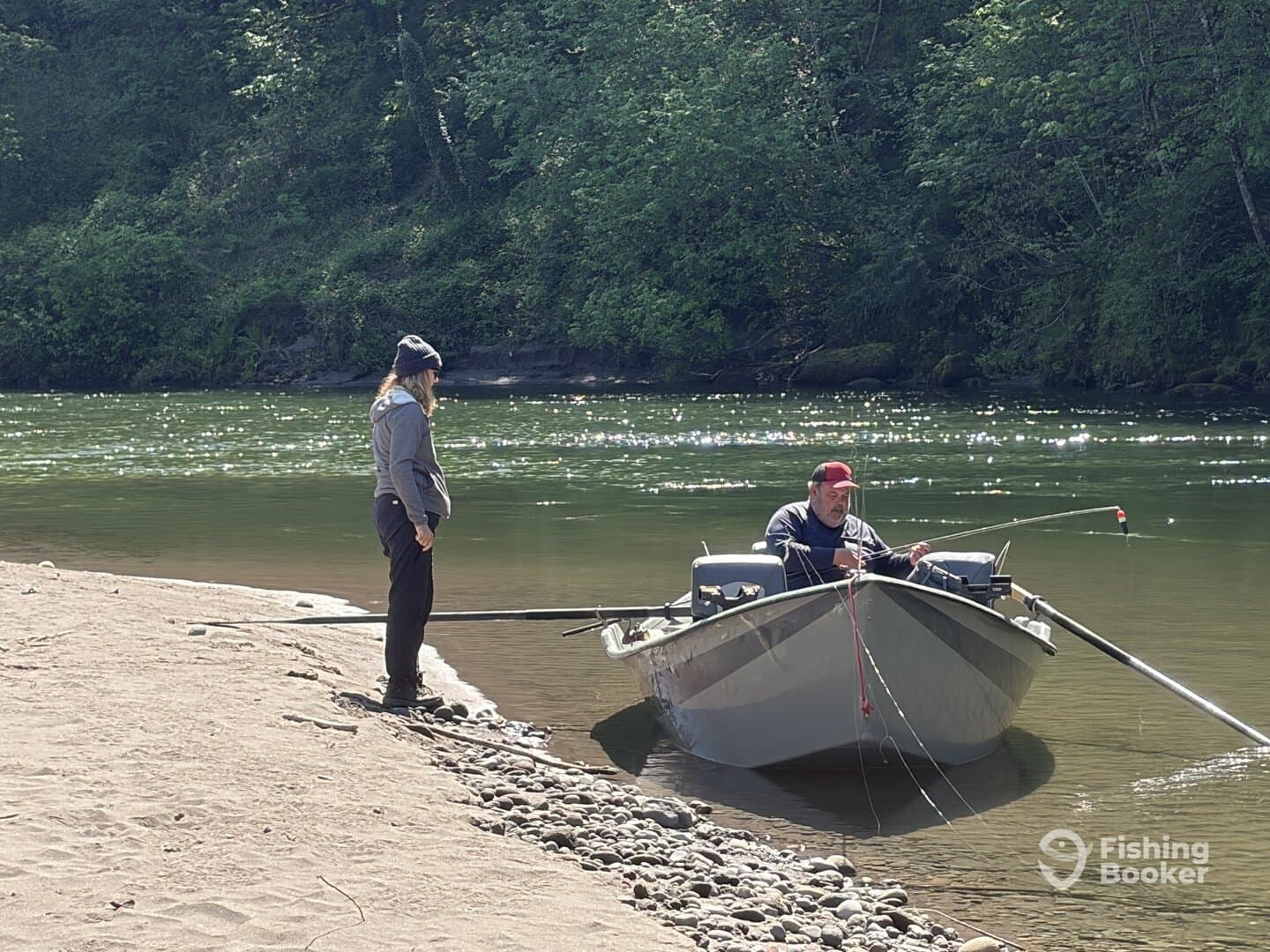 A man fishing from a small boat while a woman stands on the shore, enjoying a peaceful day on the river surrounded by lush greenery.