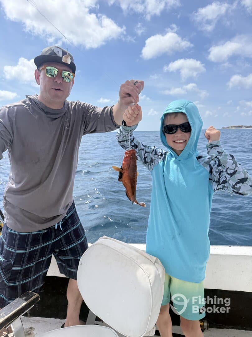 A young angler proudly holds up a Red Snapper while fishing with an adult on a boat, showcasing a fun family fishing experience.