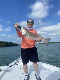Angler proudly displaying a large Redfish while standing on the bow of a boat in a scenic coastal environment.