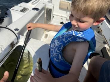 A young boy in a life jacket proudly holding a small fish while fishing on a boat.
