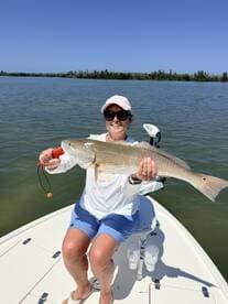 A proud angler holding a large Redfish while seated on a boat in a calm waterway, showcasing a successful day of fishing.