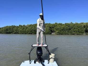 A fisherman standing on the bow of a skiff, poised to spot fish in a mangrove-lined waterway.