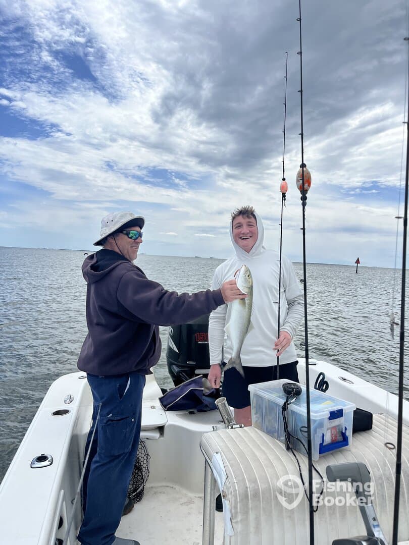 Two anglers enjoying a successful fishing trip, with one proudly displaying a caught fish on a boat in a scenic waterway.