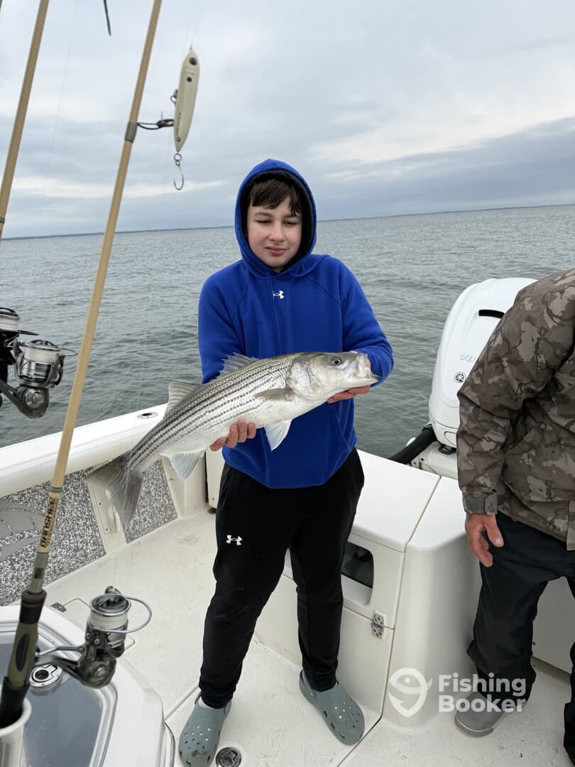 A young angler proudly displaying a large Striped Bass while fishing on a boat, showcasing a successful day on the water.