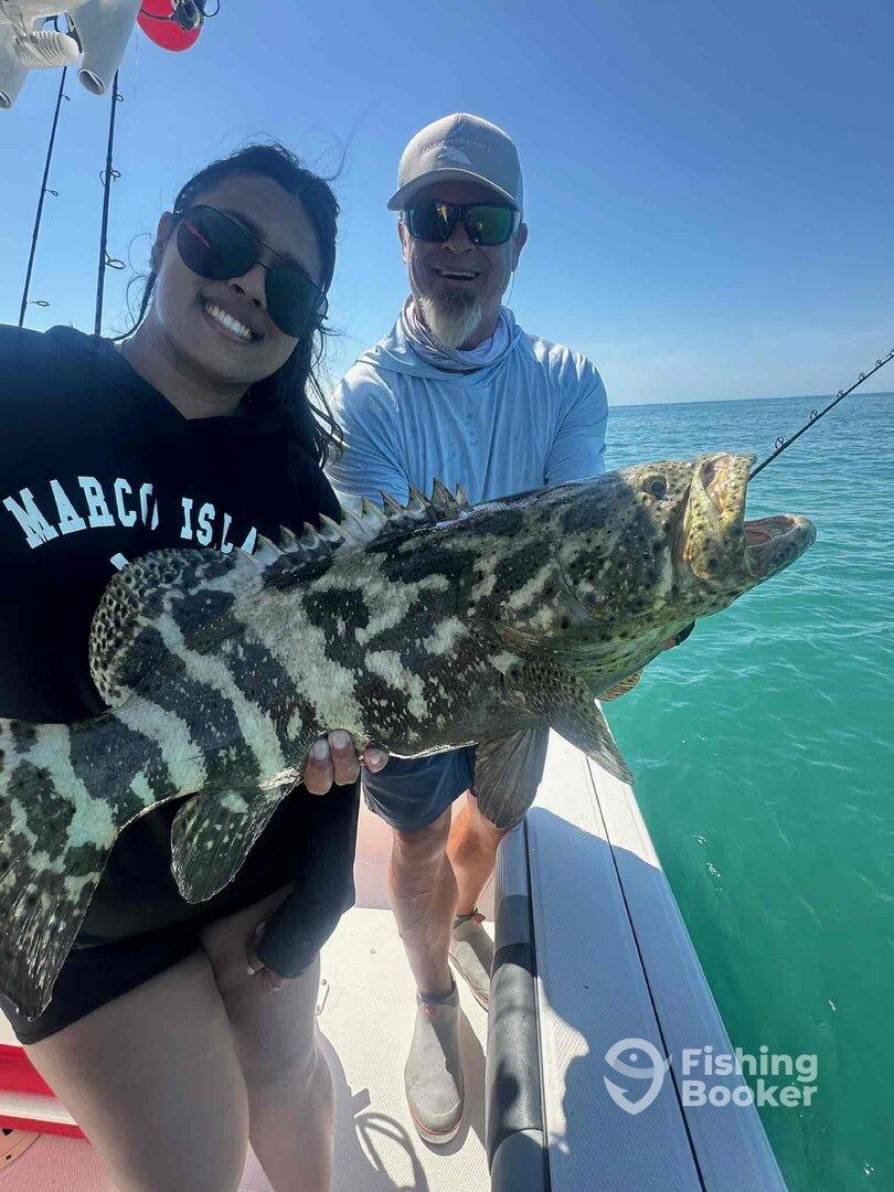 A smiling angler proudly displays a large Goliath Grouper while fishing in clear waters, showcasing a successful day on the boat.