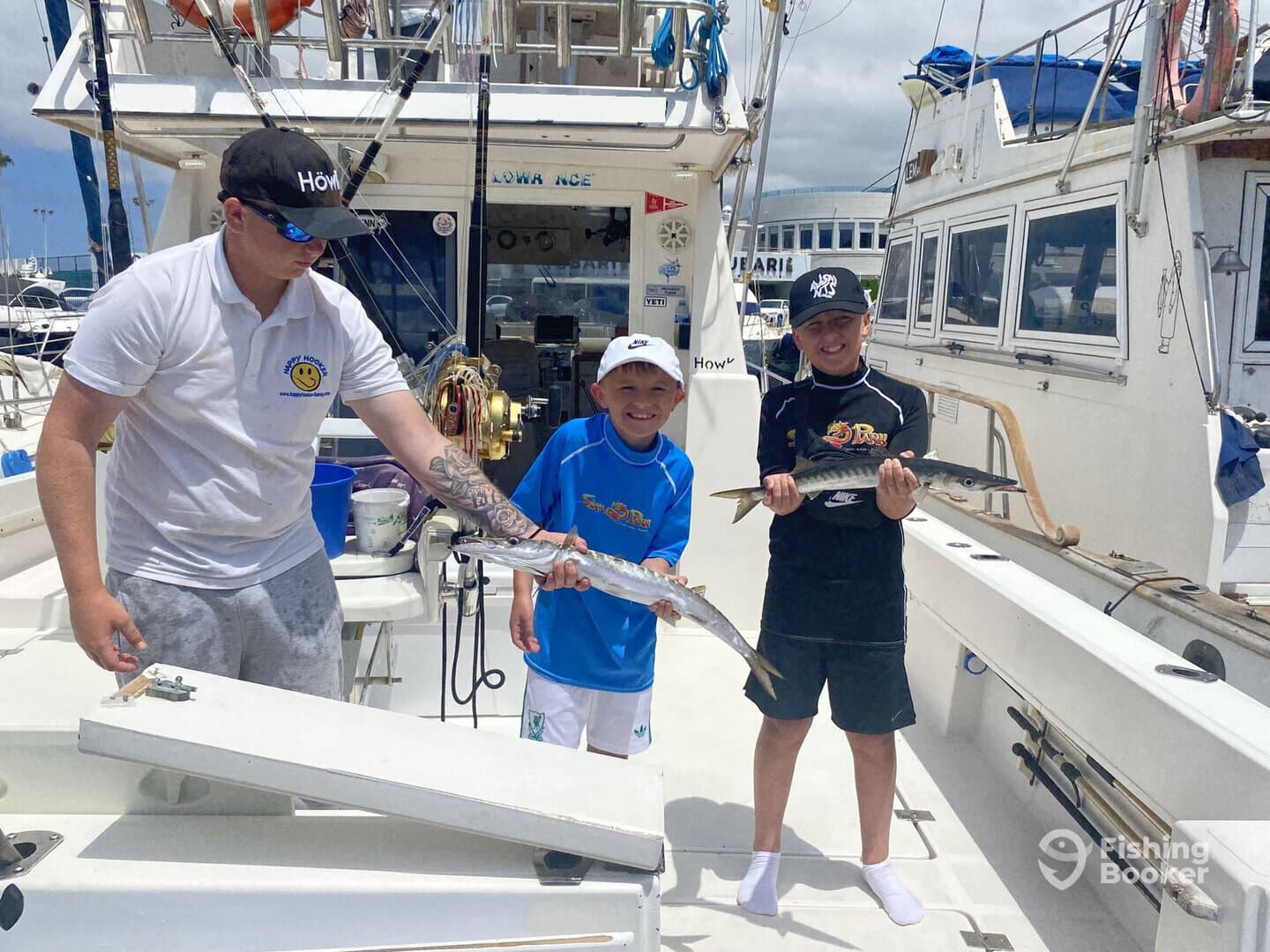 Two young anglers proudly displaying their catches of Barracuda on a fishing charter, with a crew member assisting on the boat.
