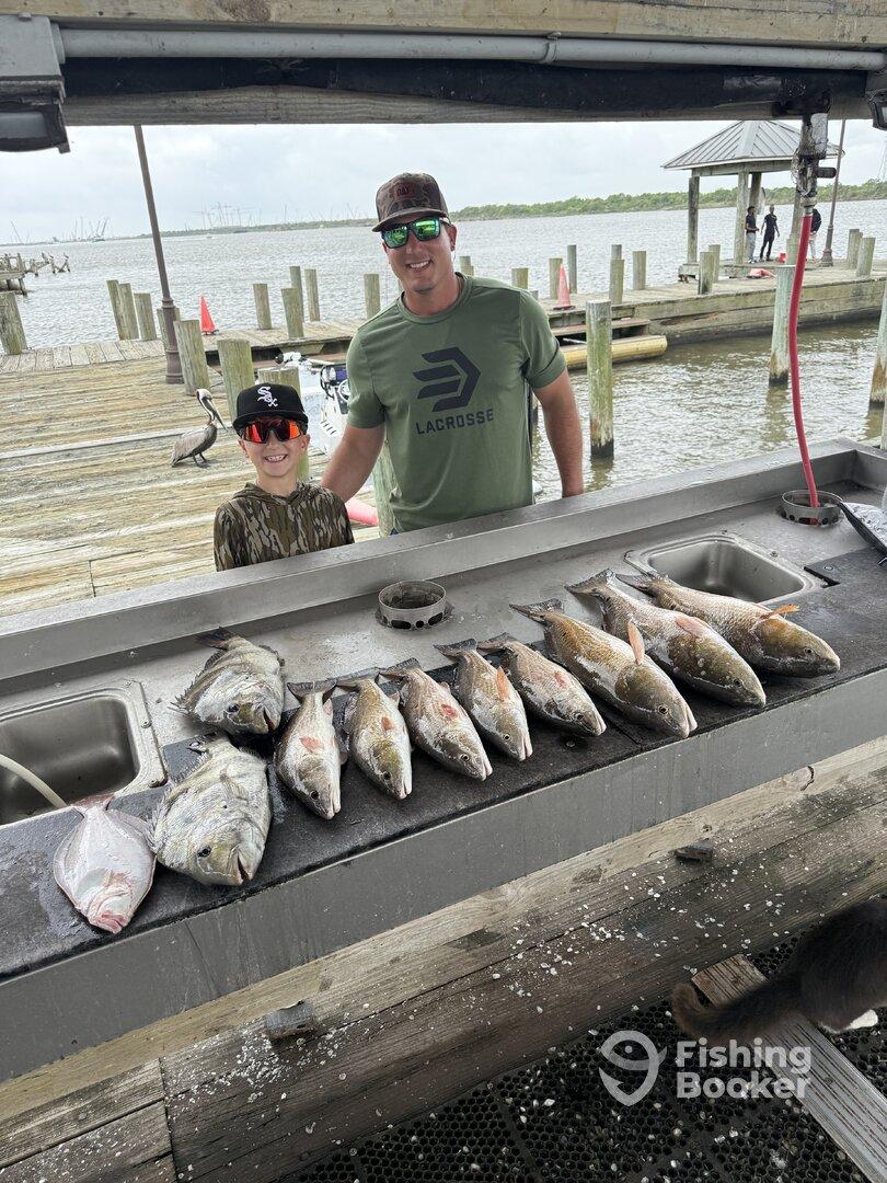 A father and son proudly stand by a cleaning station displaying a variety of fish, including Redfish and Flounder, after a successful day of fishing at the dock.