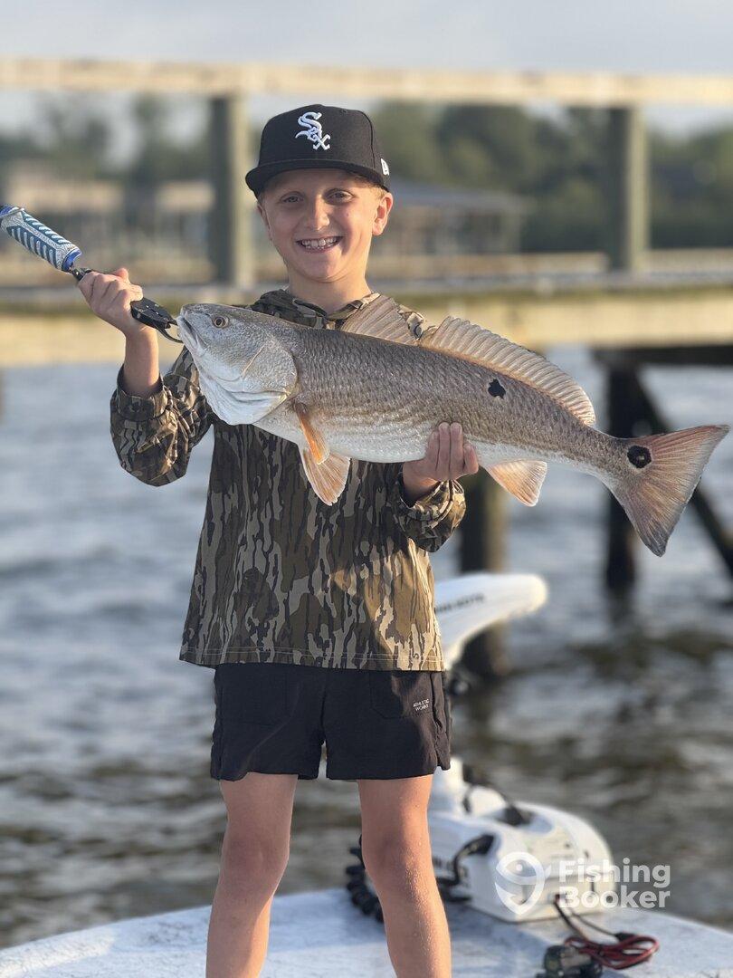 Young angler proudly holding a large Redfish while standing on a boat, showcasing a successful fishing trip.