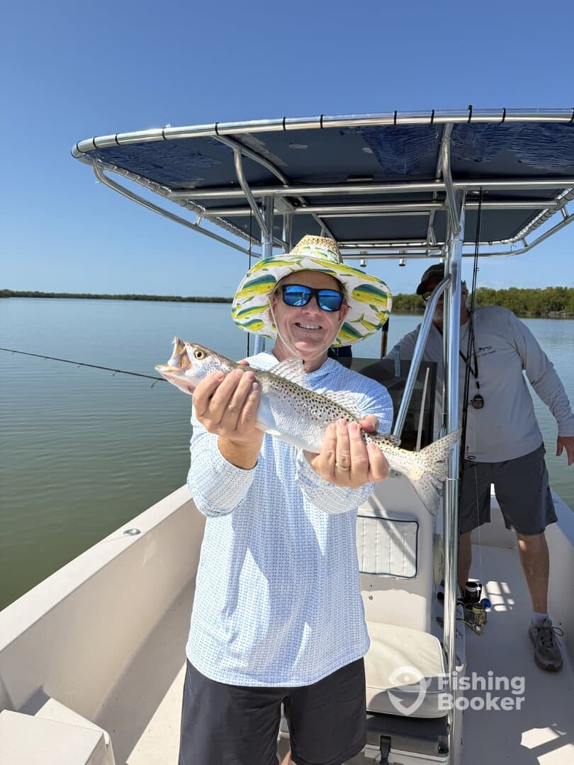 Angler proudly displaying a Speckled Trout while fishing on a sunny day in a calm waterway.