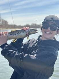 A smiling angler proudly displaying a large Brown Trout while fishing on a sunny day.
