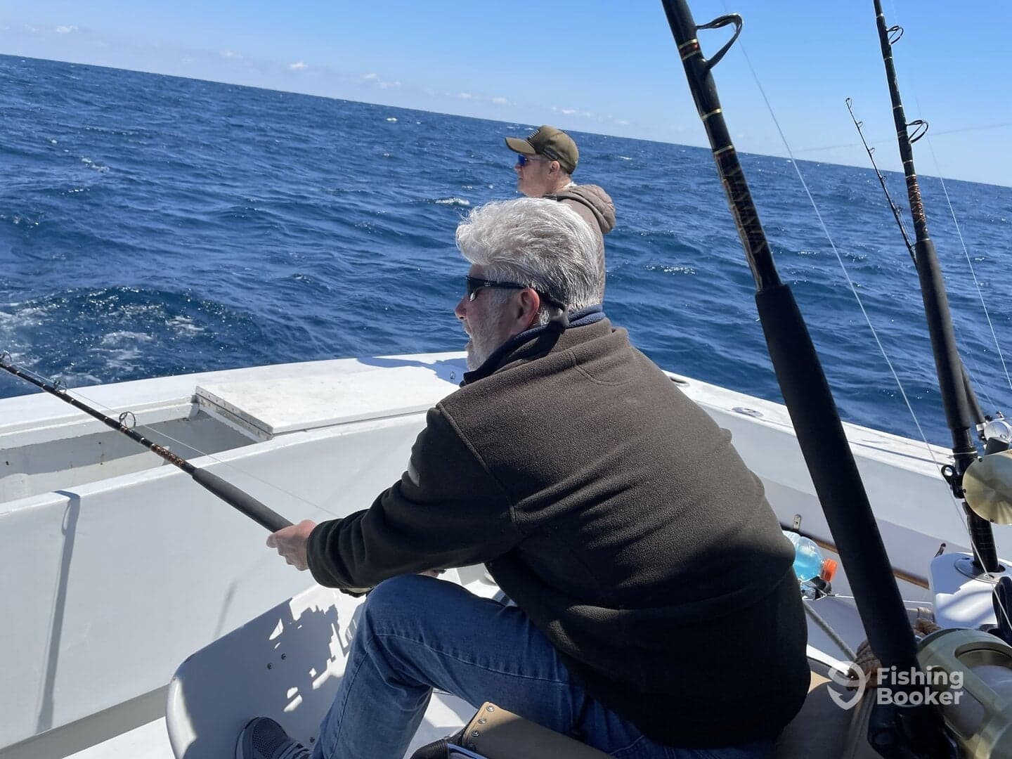Two anglers actively fishing on a boat in open water, with one reeling in a line while the other observes the ocean.
